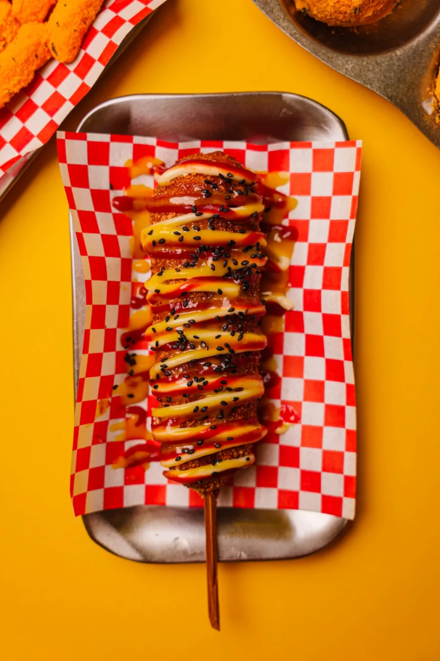 Korean corn dog con salsas y sésamo negro sobre papel de cuadros